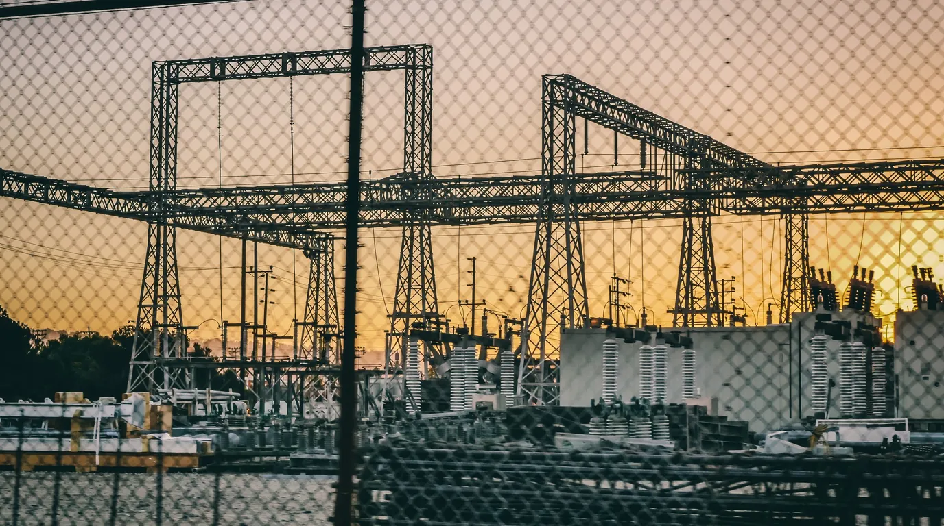 Subestação de energia vista através da cerca, com as estruturas metálicas contra o céu âmbar do fim de tarde